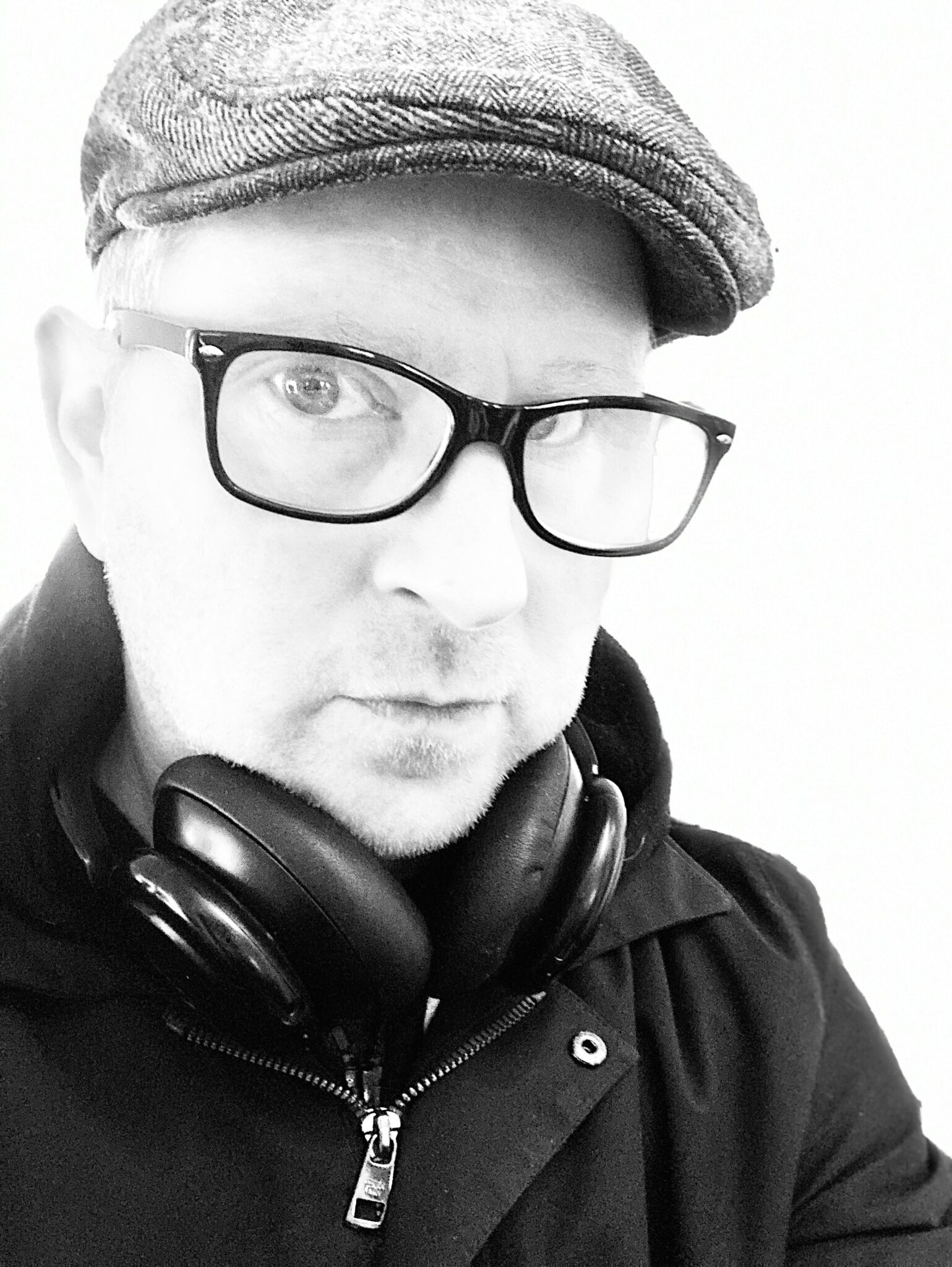 Headshot of Isaac Toast in a raincoat and newsboy cap wearing headphones around his neck and eyeglasses.